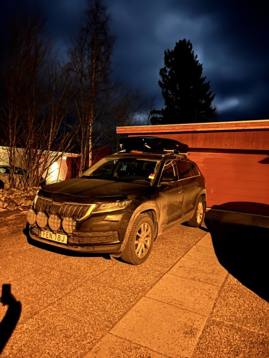 SUV parkerad nattetid, takbox, extra ljus, orange gatubelysning, mörk himmel, träd.