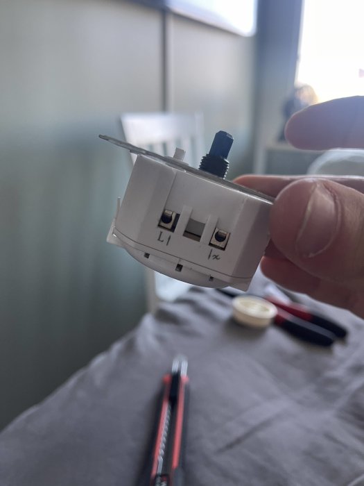 En hand håller en demonterad elektrisk väggströmbrytare framför en suddig bakgrund med verktyg.