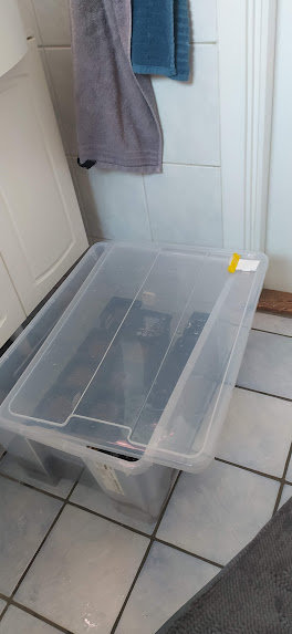 Plastlåda med lock på ett kaklat golv bredvid dörr och hängande handduk.
