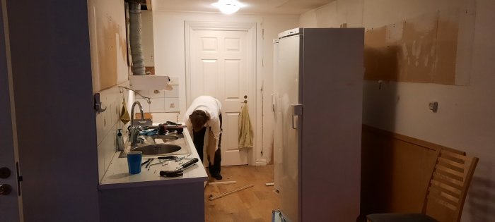 Renovering pågår i kök, person böjd över verktyg, rörigt, skåpluckor saknas, väggfläckar, ljus kylskåp, golvbrädor.