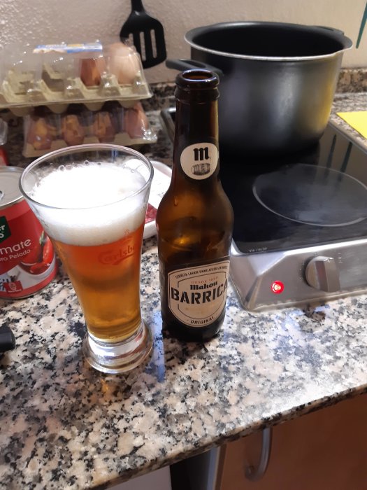 Ölglas fyllt med ljust öl, tom ölflaska, köksbänk, köksredskap, svart kastrull och spisplatta med rött ljus.