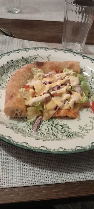 Pizza med sallad och sås på ett dekorerat porslinstallrik, bredvid ett tomt glas på ett bord.