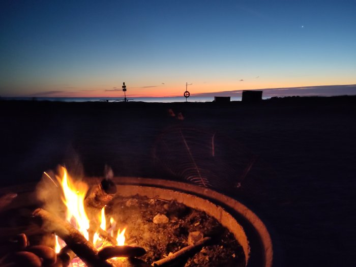 En lägereld vid skymning med färggrann himmel och en stjärna synlig. Strandnära, utomhuskväll, avkopplande miljö.