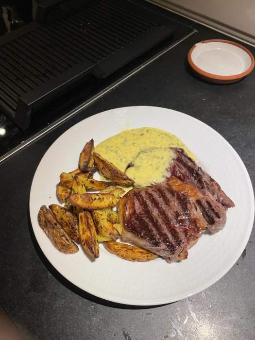 Grillad entrecote med kryddiga potatisklyftor och bearnaisesås på vit tallrik, stekhäll och liten skål i bakgrunden.