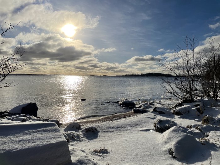 Vintersol över snötäckt strand och frusen sjö, moln, reflektioner på vattnet, blå himmel.