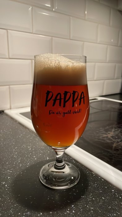 Ett ölglas fyllt med skummande dryck, text "PAPPA Du är guld värd!", på köksbänk.