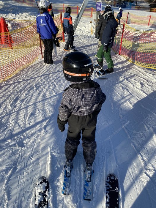 Barn i skidkläder och hjälm står på skidor vid skidliften, medan andra skidåkare väntar i kö.