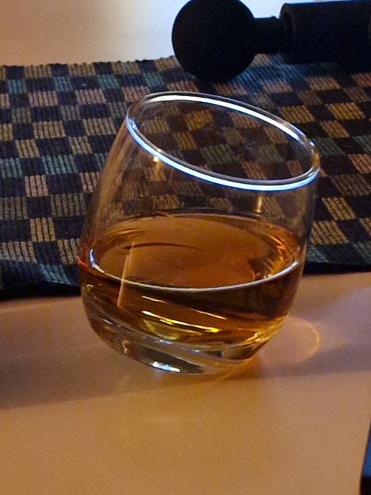 Ett snedställt whiskeyglas med gyllene vätska, på underlag, intill mörkt objekt.