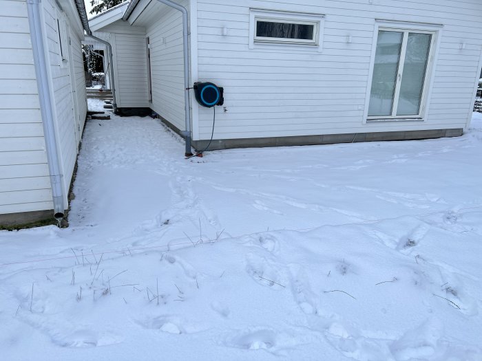 Vit villa i snölandskap, fotspår leder mot hus och blå värmepump på vägg.