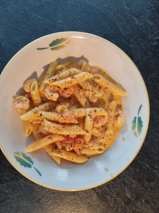 En tallrik med krämig penne-pasta och räkor, kryddad med paprika, på en blommönstrad tallrik.