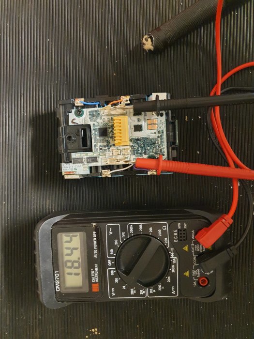 Elektroniskt kretskort kopplat till multimeter som visar 13.57 volt, möjlig felsökning eller testning på svart bakgrund.