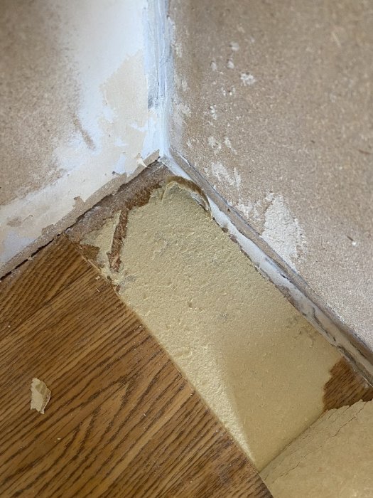 Skadad vägg och golv där tapet och färg är bortskalade, möjlig fukt- eller vattenskada synlig.