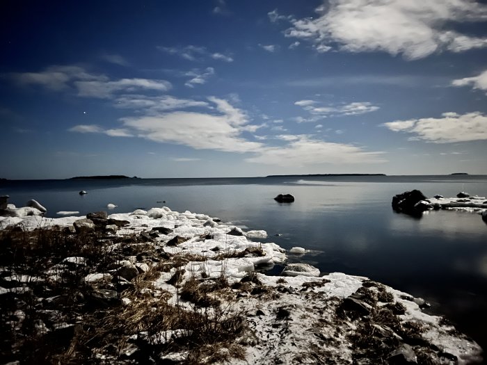 Nattlig vinterkust med snö, is, klippor och moln under stjärnhimlen.