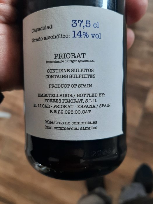 En hand håller en flaska vin med etikett som visar "PRIORAT", 14% alkoholvolym, innehåller sulfiter, produkt från Spanien.