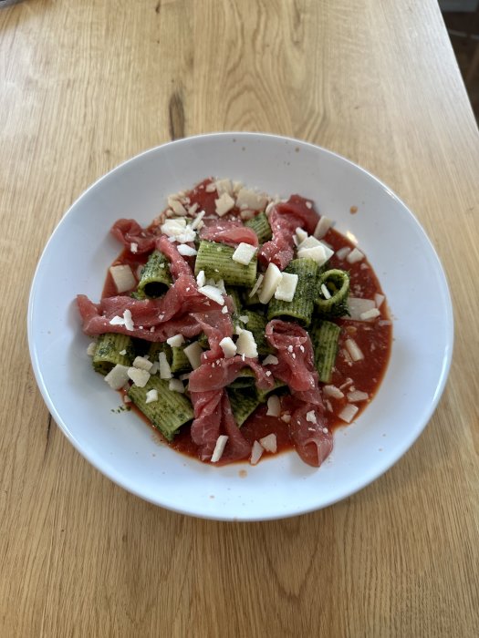 Pasta med grön pesto, skivad rå biff och riven ost i vit skål på träbord.