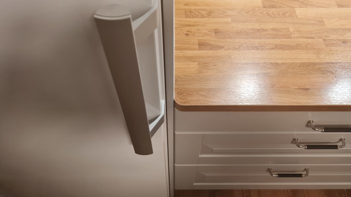 Modernt kök, trägolv, vita skåp, metallhandtag, kylskåpsdel i vänsterkant.