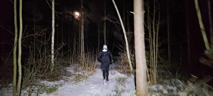 Person joggar nattetid på snötäckt stig i mörk skog med pannlampa.