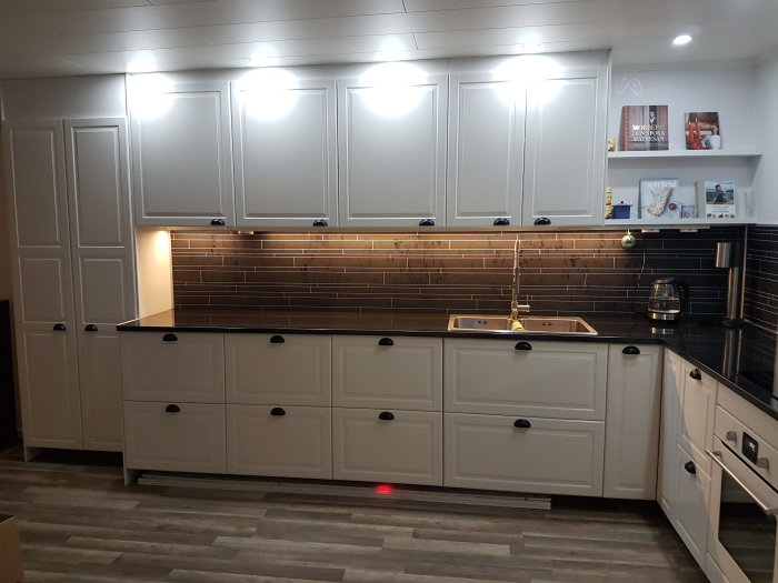 Modernt kök med vita skåp, svarta bänkskivor, mörkt kakel, rostfria apparater, öppna hyllor.