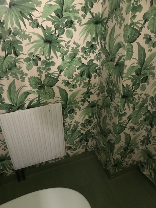 Ett rum med grön, blommig tapet, vit radiator, grön golvmatta och del av en toalett.