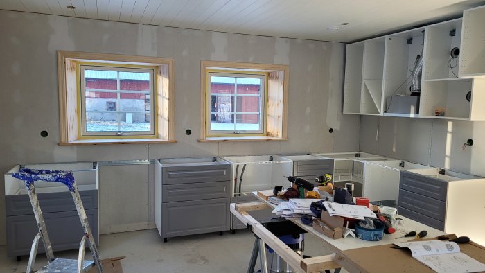 Kök under installation, oslipade väggar, fönster, verktyg, rörande utomhusvy, byggmaterial och stegar.