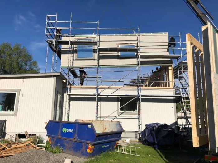 Byggnadsställning, hus under konstruktion, arbetare, byggavfall, container, trämaterial, soligt väder, utomhus.