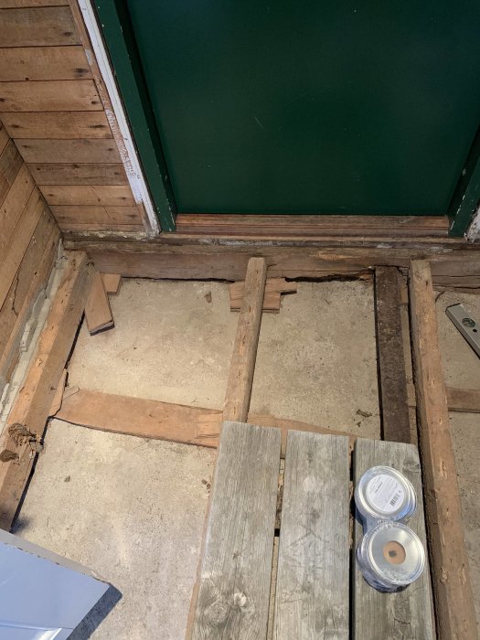 Delvis nedmonterat trägolv, betongunderlag, grönt skåp, renoveringsmaterial, oavslutat byggprojekt.
