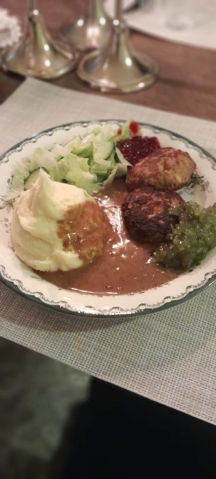 Köttbullar, potatismos, lingon, brunsås, ärtor, sallad på vit tallrik, defokuserade silverfärgade ljusstakar i bakgrunden.