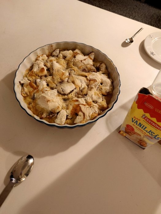 Nybakad äppelpaj i form, pudrad med florsocker, serveras med vaniljsås, sked och tallrik på bordet.