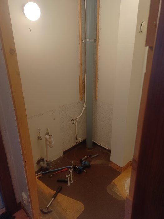 Ett tomt badrumsutrymme under renovering med rör, verktyg och golvmönster.