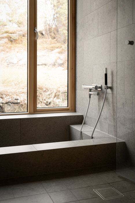 Modernt badrum, stentegel, fönster med naturutsikt, inbyggd badkar, duschmunstycke, neutrala färger.