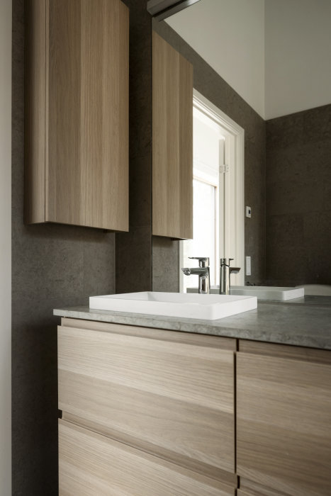 Modernt badrum, träskåp, stenbänkskiva, fyrkantig handfat, gråa toner, minimal design.