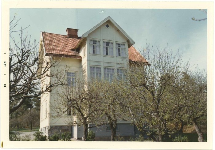 Ett traditionellt trähus med röd takpannor, omgivet av grönskande träd, klar himmel, fotografi daterat maj 1970.