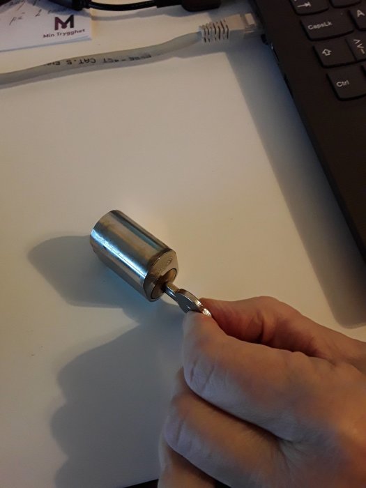 Hand håller metallstämpel över vitt papper, dator och nätverkskabel i bakgrunden, kontorstillbehör.