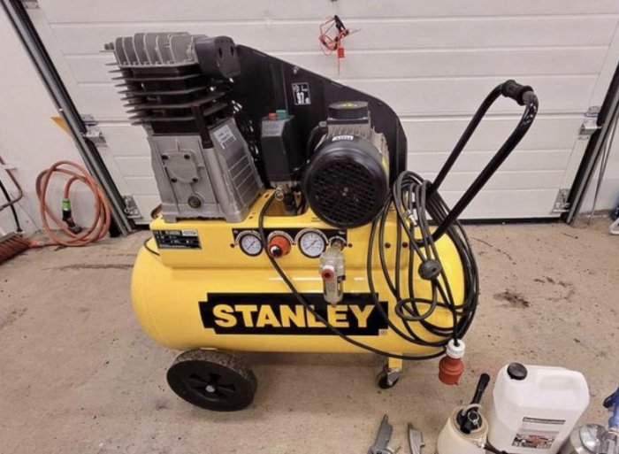 Gul luftkompressor med svart motor, mätare och slangar i ett garage, märkt med STANLEY.