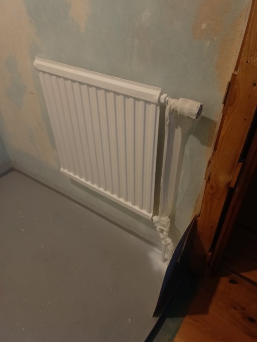 Vit radiator på en orenoverad vägg, bredvid träbalk, på ett grått golv.