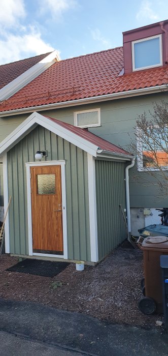 Ett grönt hus med tegelrött tak, träfärgad ytterdörr, fönster och avloppsrör, soligt väder.