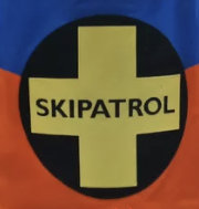 Emblem med kors, texten "SKIPATROL", antyder ski patrull eller räddningsteam, färggyllene, svart, blått, orange.