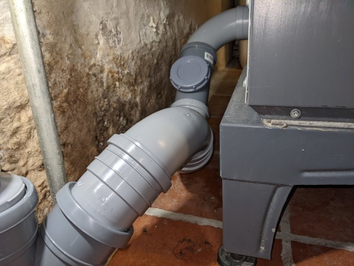 Avloppsrör i källare, råa betongväggar, rörsystem, kabel, del av möbel, tegelgolv.