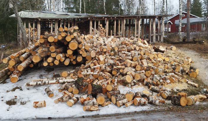 Stor hög av ved, snöfläckar, vedskjul, träd, röd stuga i bakgrunden.