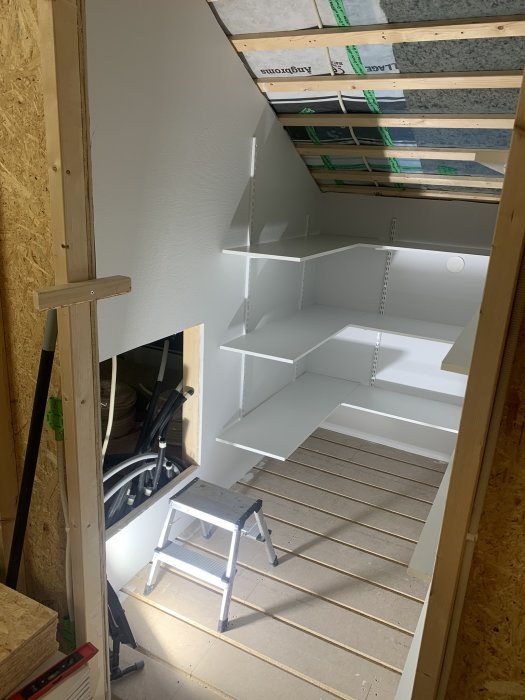 Avlutad vind med vita hyllor, stege, träreglar synliga, under konstruktion eller renovering, trägolv, isoleringsmaterial.