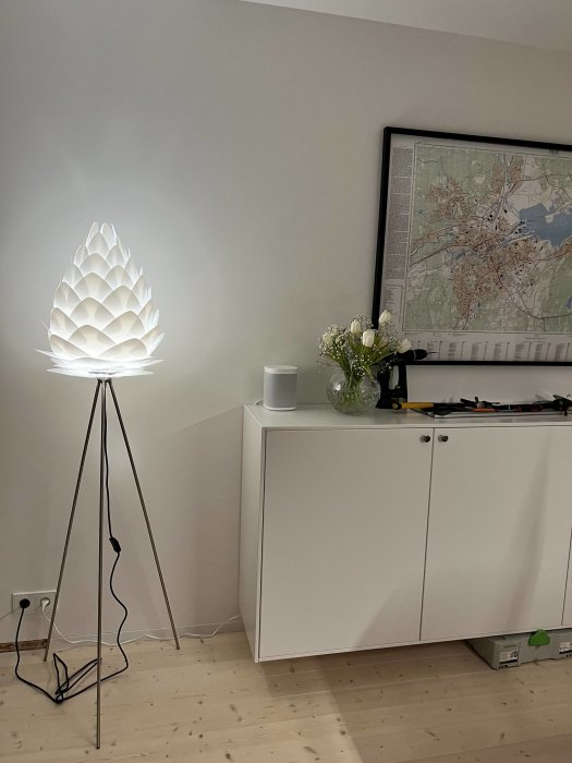 Ljus inredningsbild: designerlampa, vit skåpmöbel, karttavla på väggen, blommor, trägolv, modern stil.