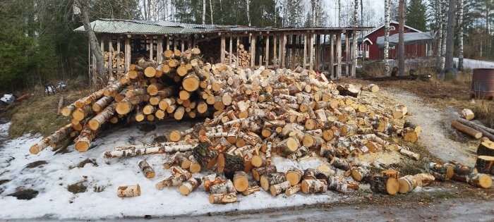 Trave av ved framför vedbod, röd stuga, snöfläckar, barrträd, landlig miljö.