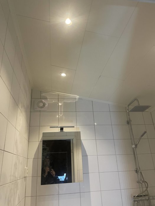 Ett badrum med vita kakelväggar, taklampor, dusch, spegelskåp och person som fotograferar spegelbilden.
