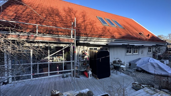 Takarbete på ett hus med tegeltak, ställning, trädgårdsmöbler täckta, soldränkt terrass, vindstilla dag.