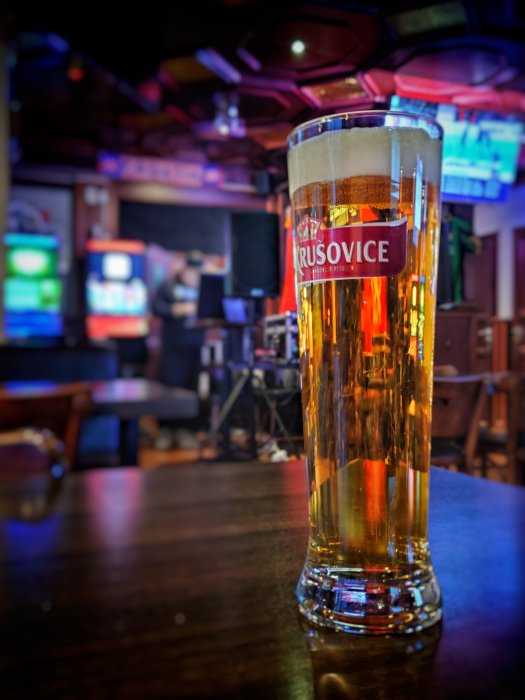 Ölglas fyllt med öl på bar, suddig bakgrund med TV och person. Färgrik, djup skärpedjup, avslappnad atmosfär.