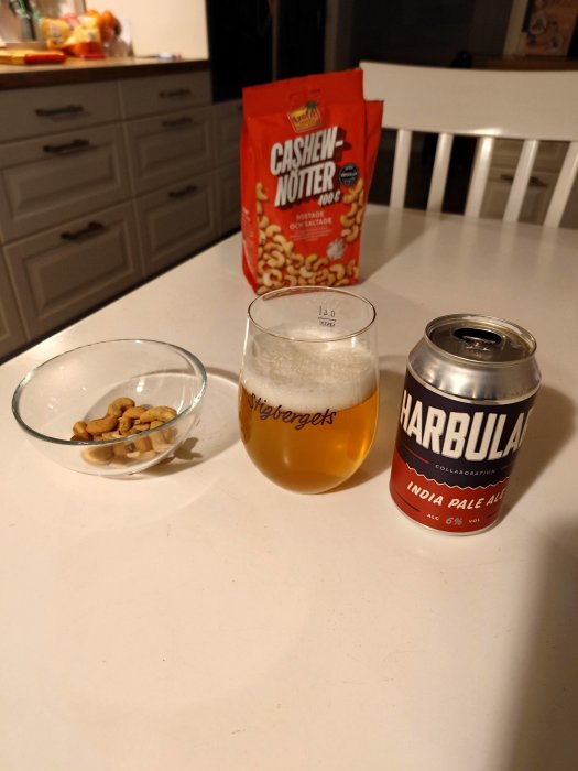 Ett glas öl, ölburk, rostade och saltade cashewnötter på ett köksbord.