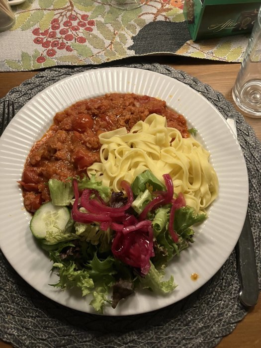 En tallrik med pasta, köttsås och sallad på ett matbord, med servett och glas bredvid.