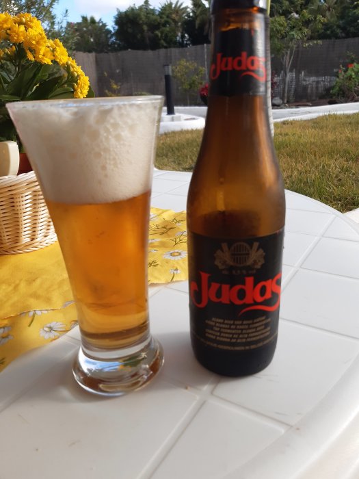 En ölflaska och ölglas på ett bord utomhus, med grönska och blommor i bakgrunden.