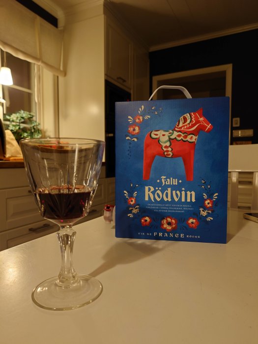 Ett glas rödvin på bänk, Falu Rödvin-påse med hästmotiv, inomhuskök, mysig belysning, kvällstid.