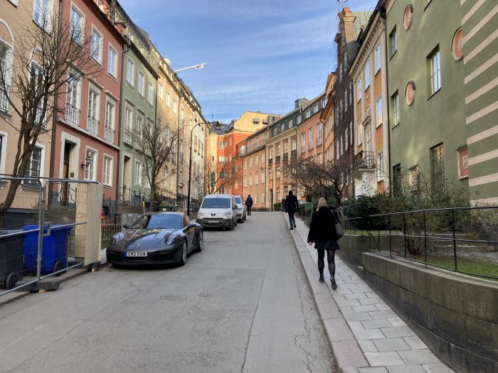 Stadsgata med folk, färgglada byggnader, parkerade bilar, blå himmel, utan identifikation av personer eller fordon.